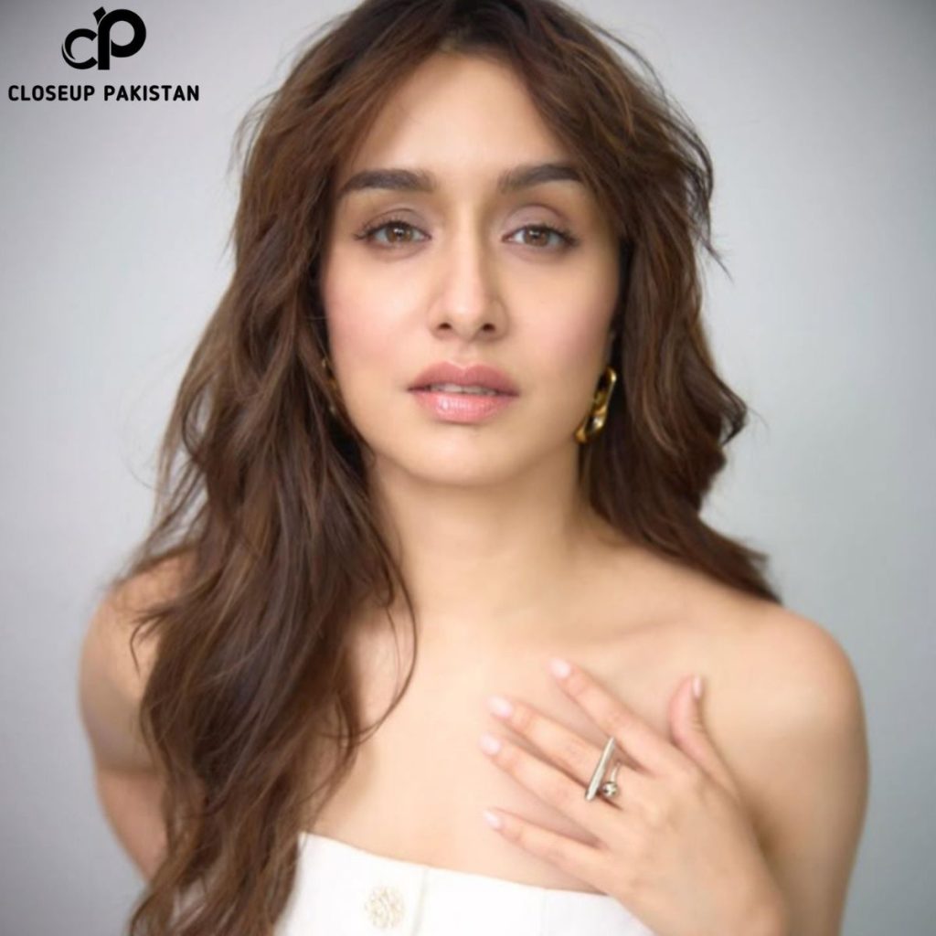A woman with long wavy brown hair poses against a light background, wearing a strapless white outfit, gold earrings, and a ring on her left hand, which rests near her collarbone.