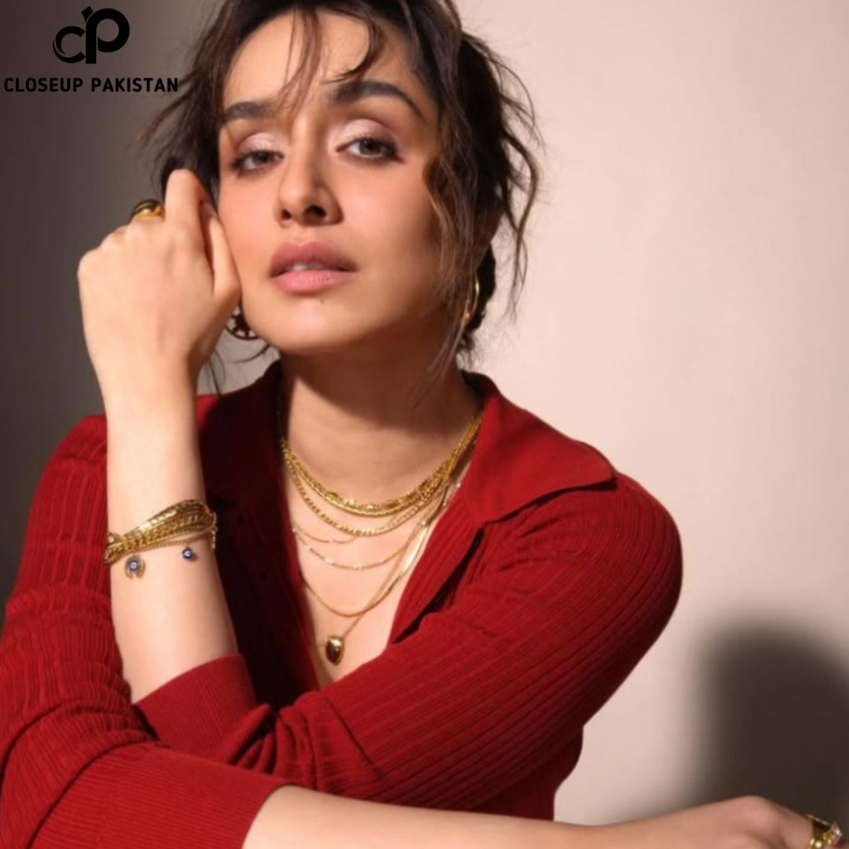 A woman with tied-back hair and loose strands framing her face poses against a neutral background, wearing a red ribbed top, layered gold necklaces, and gold bracelets with a thoughtful expression.
