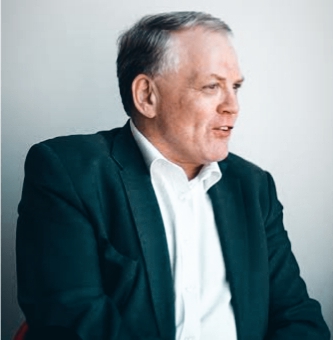 Man in a dark green blazer and white shirt, sitting and speaking while facing right, against a light gray background.