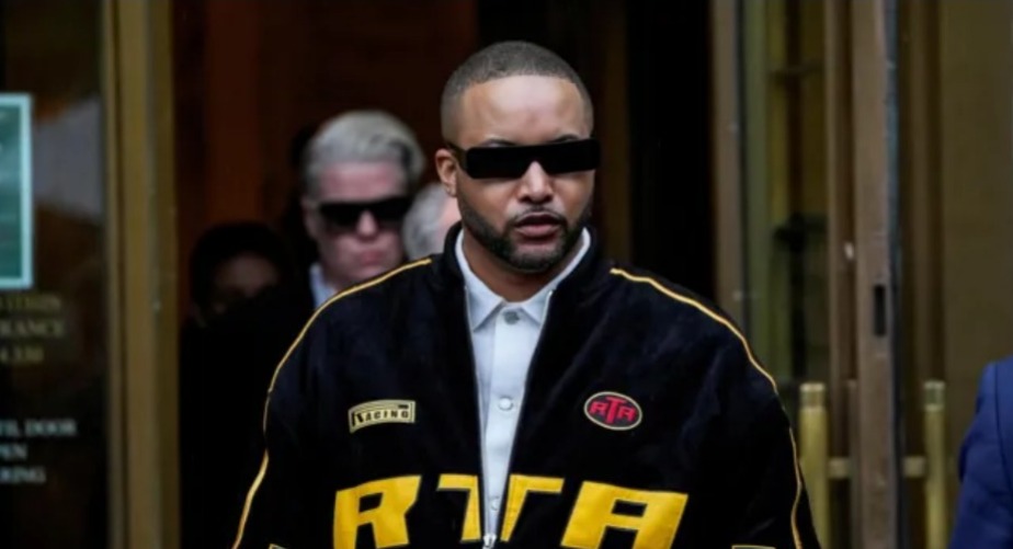 A man wearing black futuristic-style sunglasses and a dark jacket with yellow "RTA" lettering walks confidently out of a building. He has a trimmed beard and a closely shaved haircut. Other people, also wearing dark glasses and formal clothing, follow him in the background. The setting appears to be outside a courthouse or official building with golden doors.