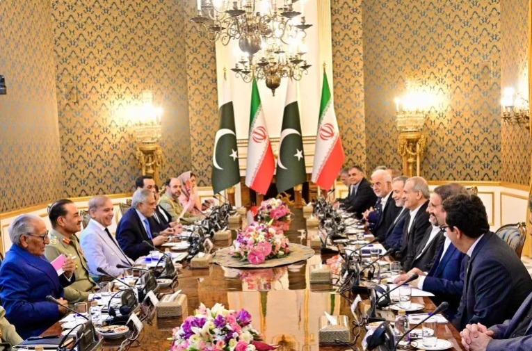 Delegations from Pakistan and Iran sit across from each other at a large ornate conference table in a formal diplomatic meeting. Behind them are flags of both countries – Pakistan and Iran. The setting is elegant, with chandeliers, patterned wallpaper, and flower arrangements on the table.