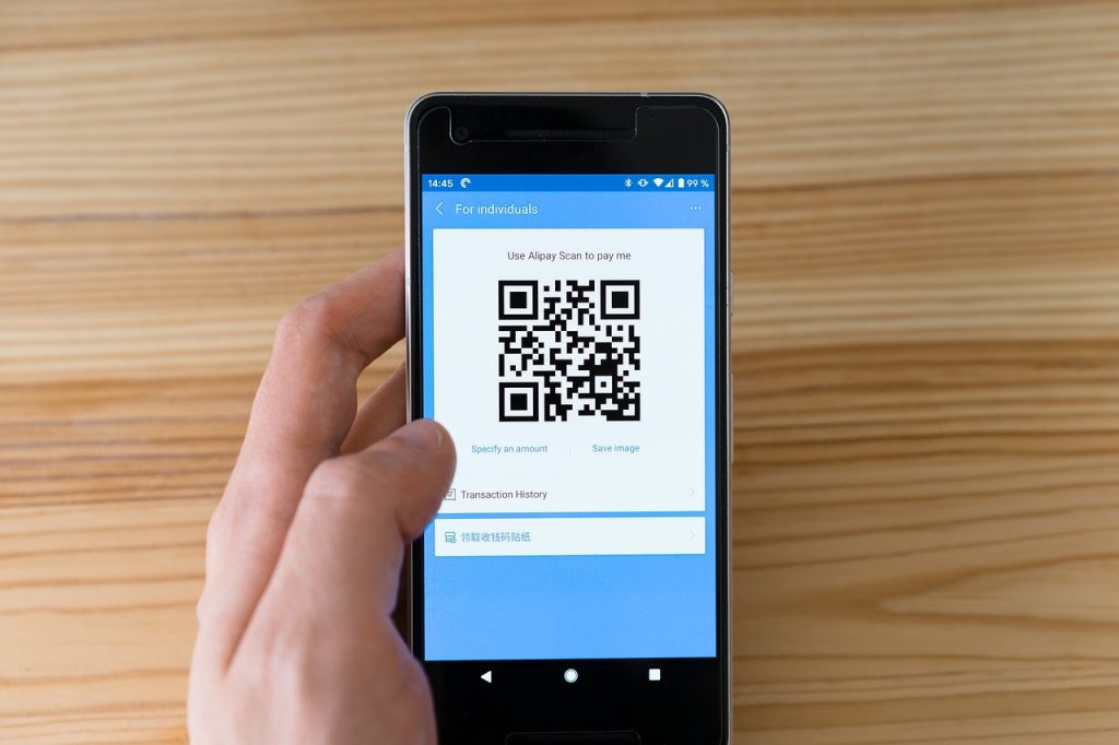 A person holding a smartphone displaying a QR code on the Alipay app screen, with options for specifying an amount and viewing transaction history.