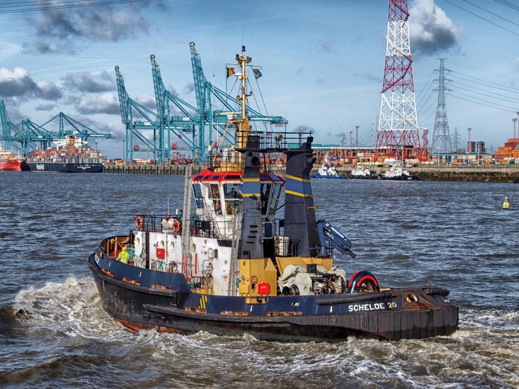 A tugboat named "SCHELDE 20" navigates through the water in an industrial port. In the background, large container cranes, cargo ships loaded with containers, and tall communication towers dominate the scene. The sky is partly cloudy with high-voltage power lines visible overhead, emphasizing the industrial setting. The water is slightly choppy, and crew members are visible on the deck of the tugboat.