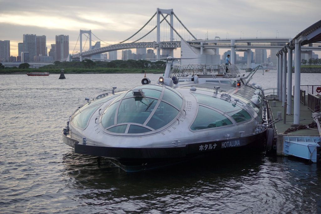 Futuristic-looking silver passenger boat named "Hotaluna" docked at a pier in Tokyo Bay, with the Rainbow Bridge and city skyline in the background during sunset.