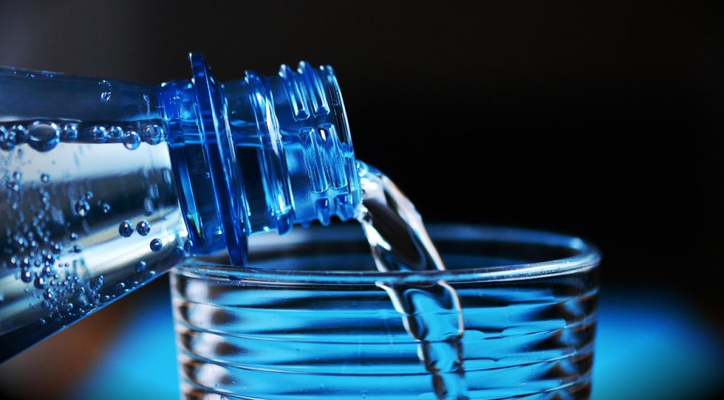 A close-up image of water being poured from a blue plastic bottle into a transparent ribbed glass. The background is dark, highlighting the clarity of the water and the texture of the bottle and glass.