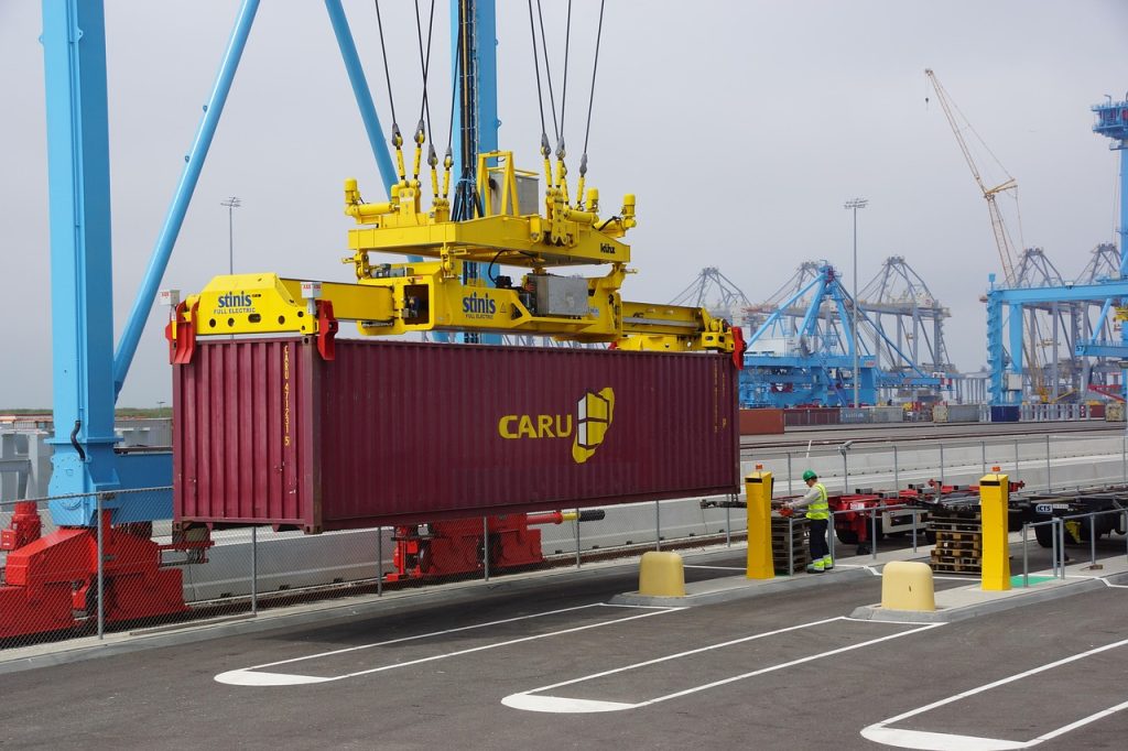 A crane lifts a CARU container at a port, with a worker nearby and blue cranes in the background.