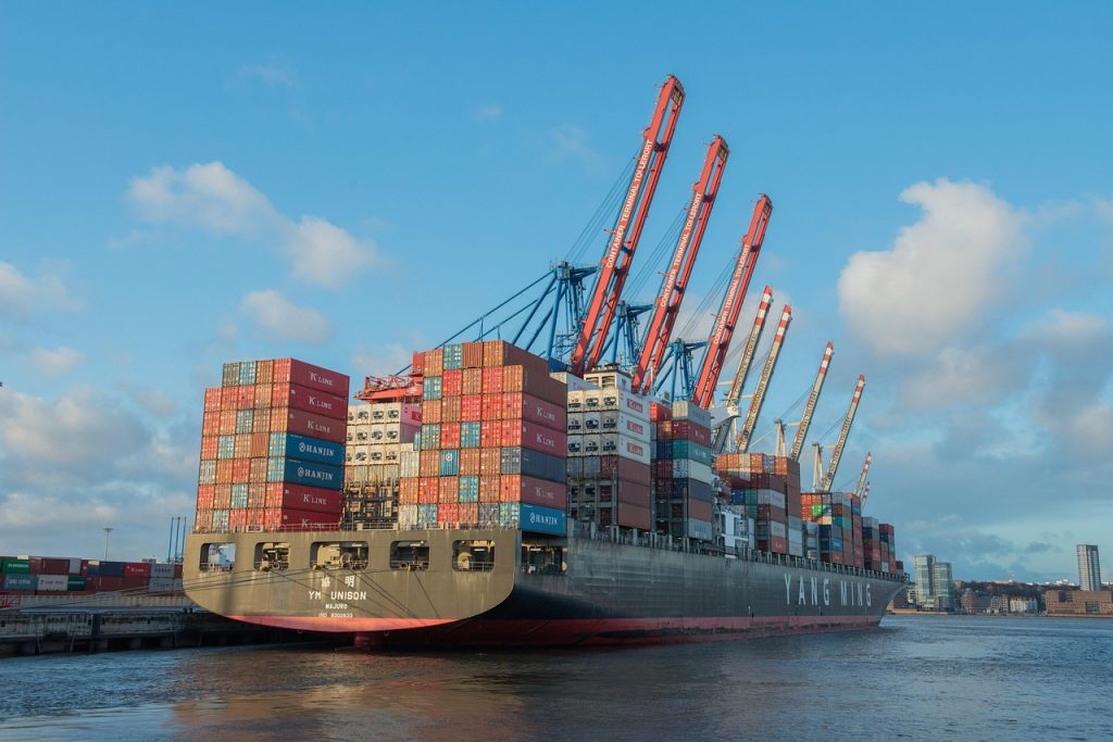 A large cargo ship is docked at a port, with several cranes visible in the background. The ship is loaded with numerous shipping containers of various colors, including red, blue, and white. The sky above is a bright blue with some clouds.