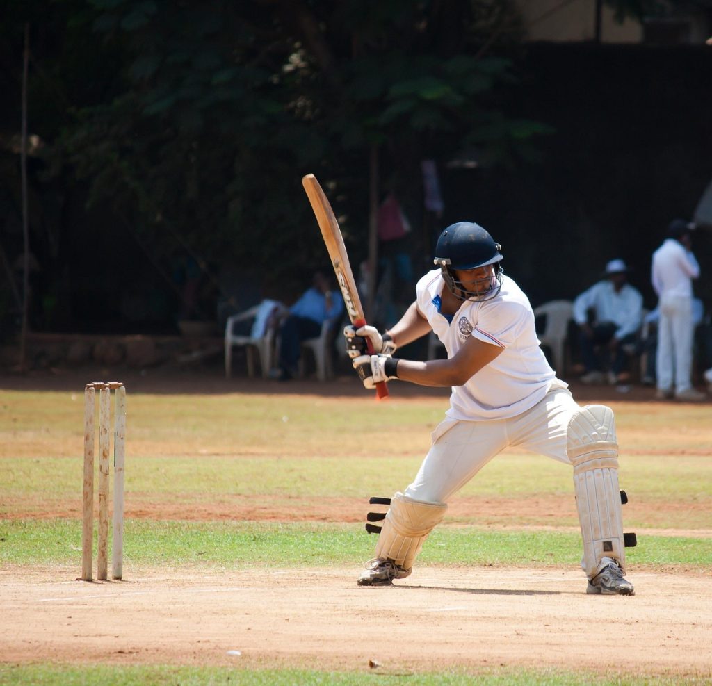 Cricketer in white uniform, batting on a cricket pitch with spectators in the background.