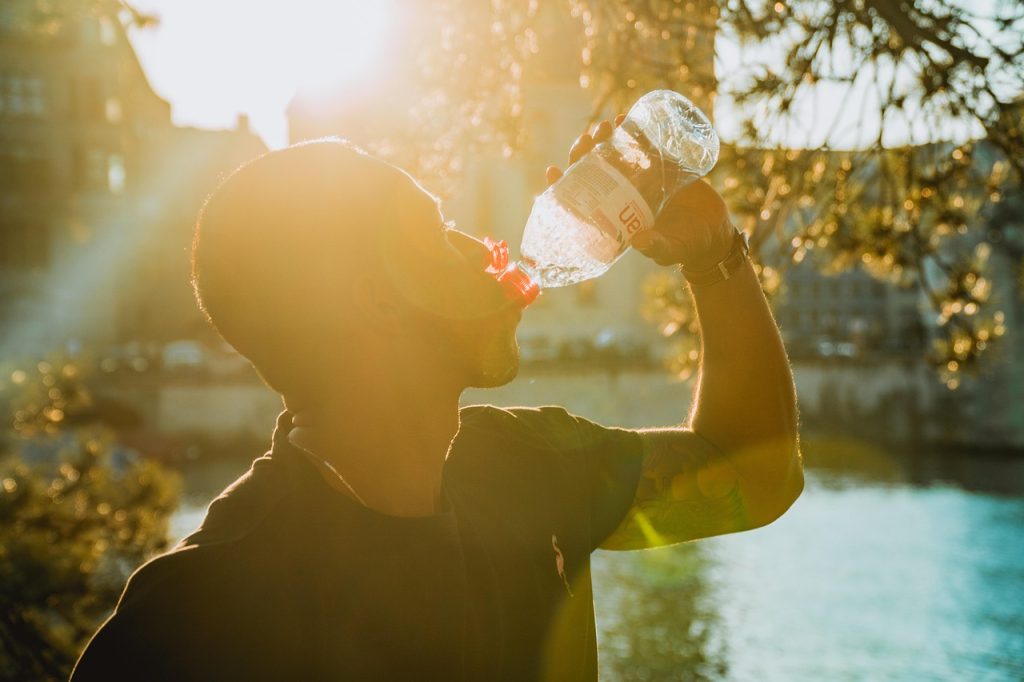 A man is silhouetted by the sun as he drinks from a plastic water bottle outdoors near a body of water. The sunlight creates a warm, golden glow around him, and trees and buildings are visible in the background.