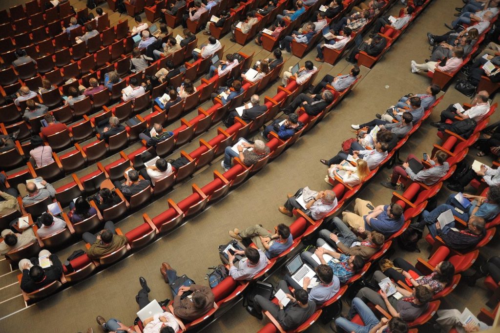 **Alt Text:**
Large audience seated in red chairs at a conference or seminar in an auditorium, attentively listening, with many using laptops, notebooks, and mobile devices.
