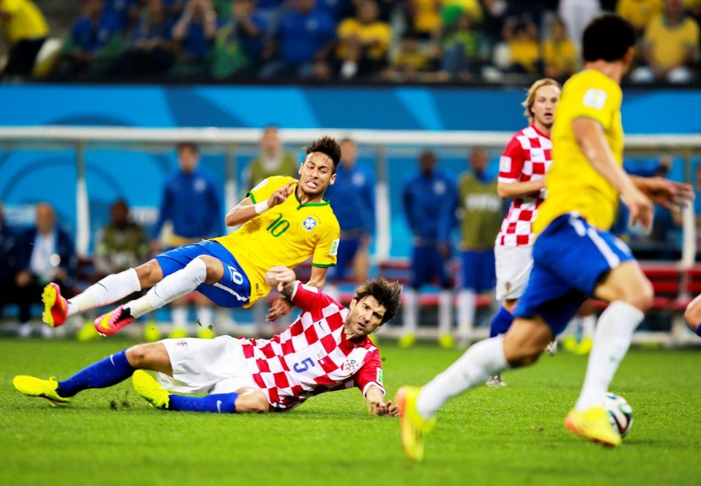 A dynamic moment from a football match, where a player in a yellow jersey (Brazil) wearing number 10 is tackled by an opponent in a red-and-white checkered jersey (Croatia). The action takes place on a green field under bright lights, with a crowd in the background adding to the intense atmosphere of the game.