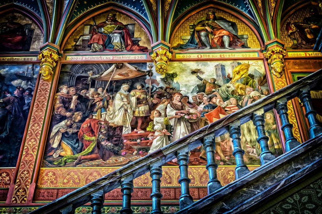 A richly detailed mural inside a historic building depicts medieval scenes of kings, knights, and clergy in ceremonial and regal settings. The artwork features vibrant colors, elaborate costumes, and dramatic storytelling. A decorative stone staircase cuts across the foreground, leading up past the wall adorned with these intricate frescoes. The scene is filled with historical symbolism and ornate architectural elements.