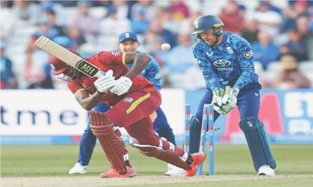 A cricket player in a red uniform is batting, with a wicketkeeper in blue behind him. The batsman is swinging his bat at an incoming ball, while the wicketkeeper is crouched and ready to catch it. The background shows a crowd of spectators watching the game.