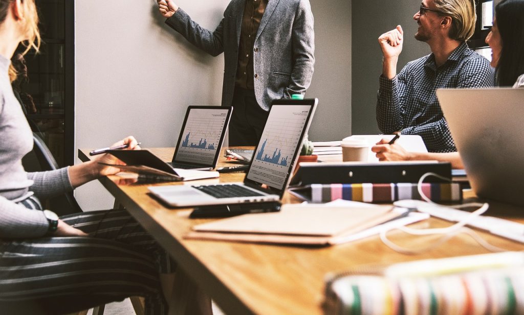 **Alt Text (Shortened):** Business team in a meeting with laptops showing graphs, one person presenting at a whiteboard.