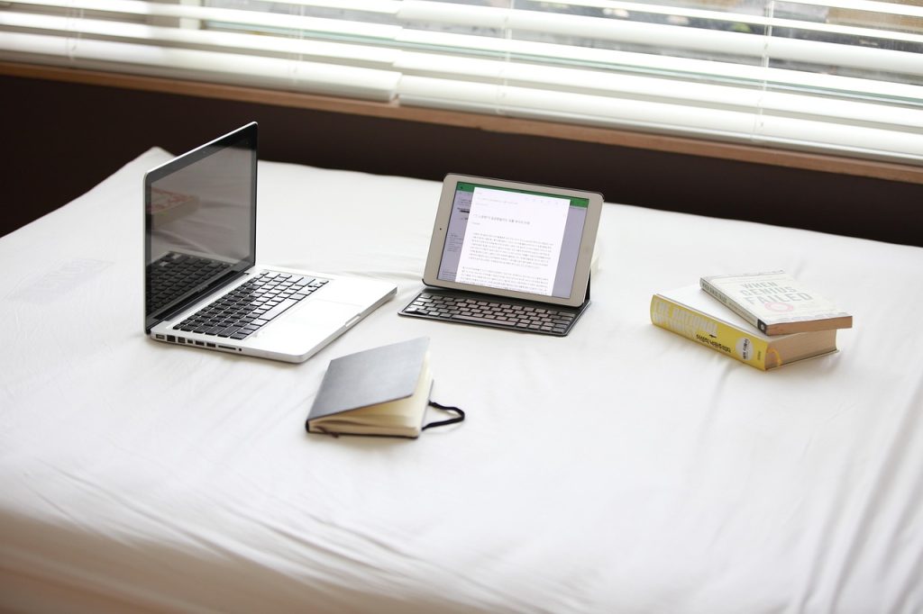 A home workspace setup on a white bed, featuring an open laptop, a tablet with a keyboard, a black notebook, and two books titled "The Rational Optimist" and "When Genius Failed," with blinds partially open in the background.