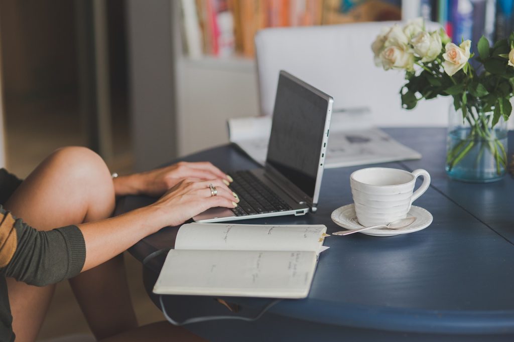 **Alt Text (Shortened):**
Person typing on a laptop at a table with a notebook, coffee cup, and vase of flowers.
