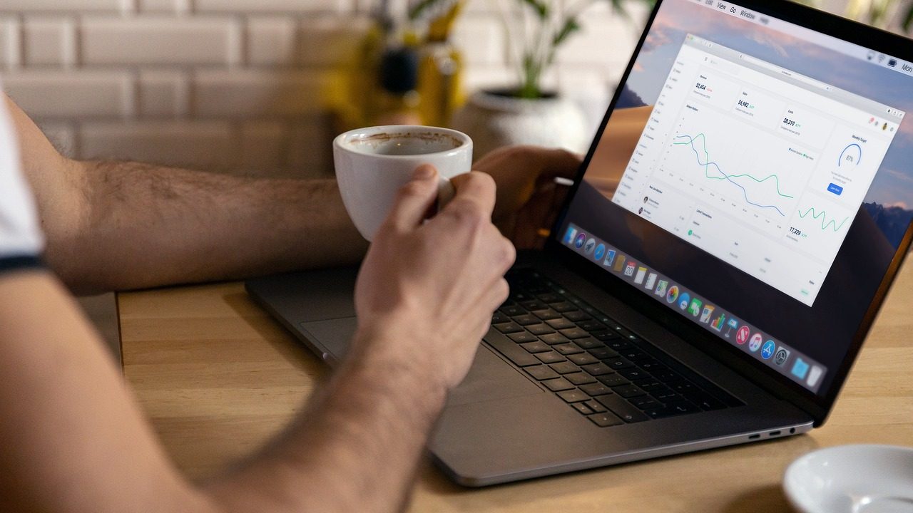 A person holding a cup of coffee while working on a laptop displaying data analytics and charts, seated at a wooden table with a white plate in the foreground and decorative plants in the background.