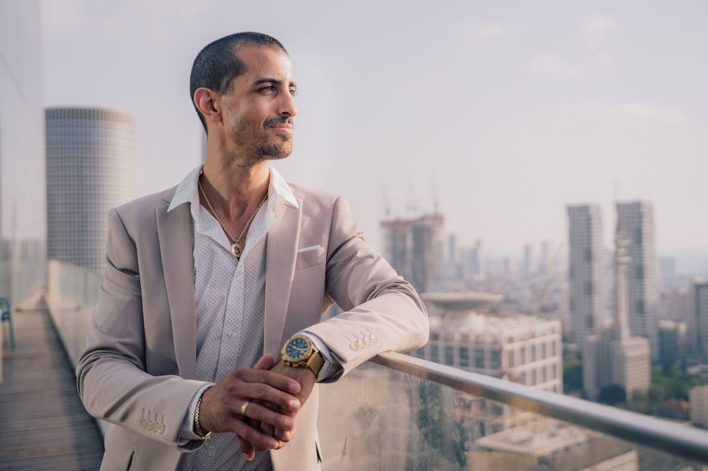  A man dressed in a light beige suit with a white shirt and gold accessories, including a watch and necklace, stands confidently on a rooftop balcony overlooking a modern city skyline with high-rise buildings and cranes in the distance.