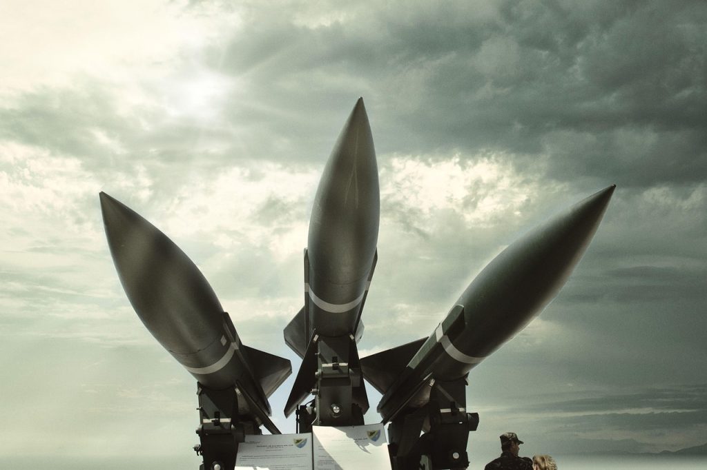 **Alt text:** Three large military missiles mounted on launch platforms, pointed skyward under a cloudy sky with two people observing below.
