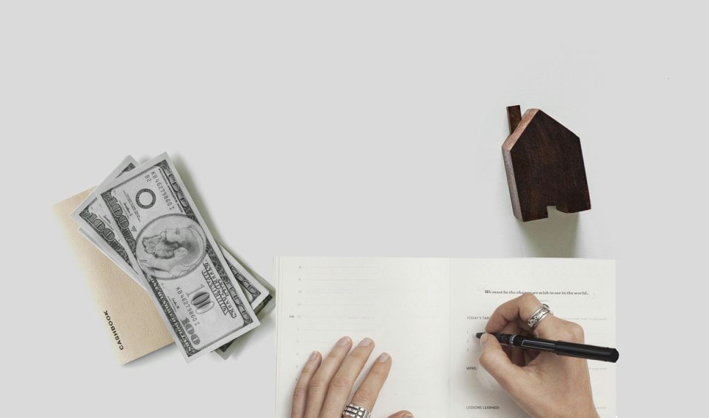  "A flat lay of a hand writing in a journal or planner. To the left are a stack of US dollar bills on a cashbook, and to the right is a small wooden house figurine."