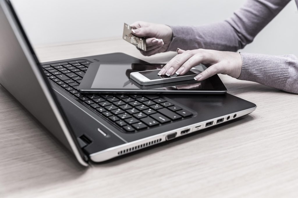 A person making an online payment using a credit card while interacting with a smartphone placed on top of a tablet and laptop.