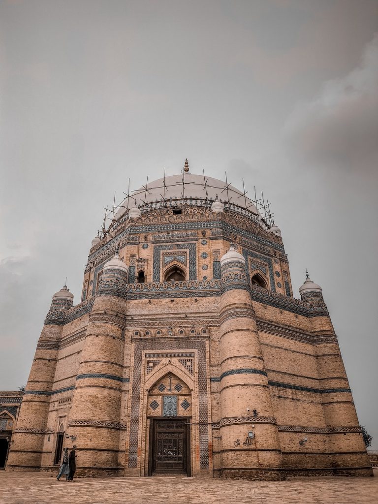 **Alt Text:**
Historic brick mausoleum with intricate blue tilework and a large white dome, located in Pakistan.
