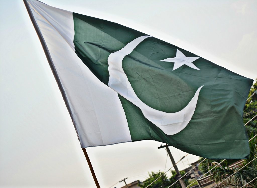 A large flag of Pakistan waving in the wind, photographed from a low angle against a light, overcast sky. The flag features a white vertical stripe on the left, with a dark green field on the right containing a white crescent moon and a five-pointed star. Electric poles and greenery can be seen in the blurred background.