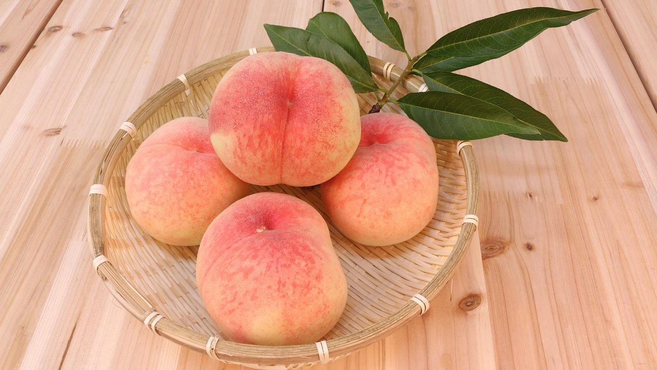 Four ripe white peaches with green leaves in a woven basket on a wooden surface.