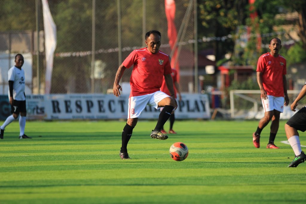 The image shows a football match in progress. A player in a red jersey and white shorts, with the number 8, is actively controlling the ball on a well-maintained green field. Other players, wearing both red and white jerseys, are visible in the background along with banners promoting sportsmanship. The setting appears to be an organized outdoor match in sunny conditions.