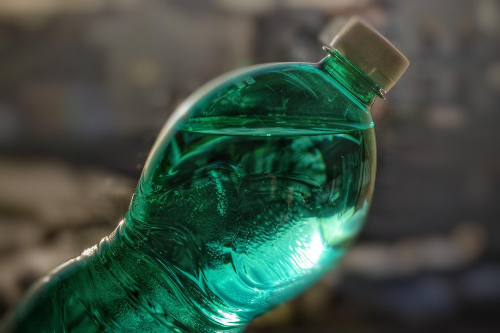 A close-up image of a green-tinted plastic water bottle filled with liquid, shown at an angle with a white cap. The background is blurred, highlighting the texture and reflections on the bottle's surface.