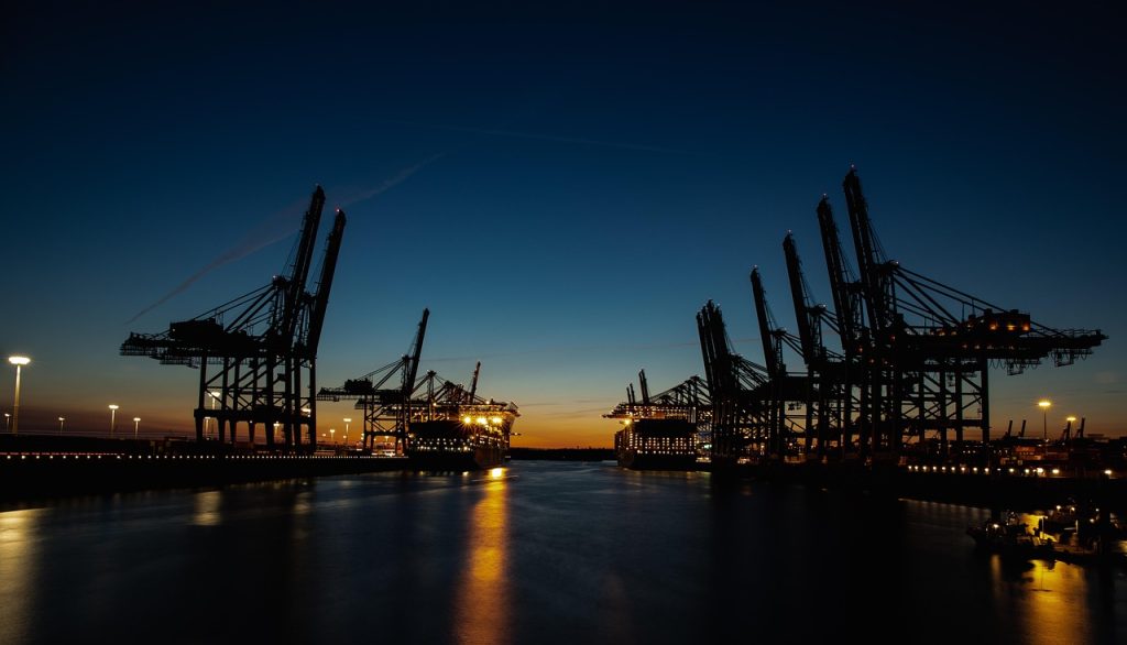 A nighttime view of a shipping port with large cargo cranes silhouetted against a twilight sky. The sky transitions from deep blue at the top to orange near the horizon. The port is lit with artificial lights, and the calm water reflects both the lights and the dark structures of the cranes, creating a serene industrial scene.