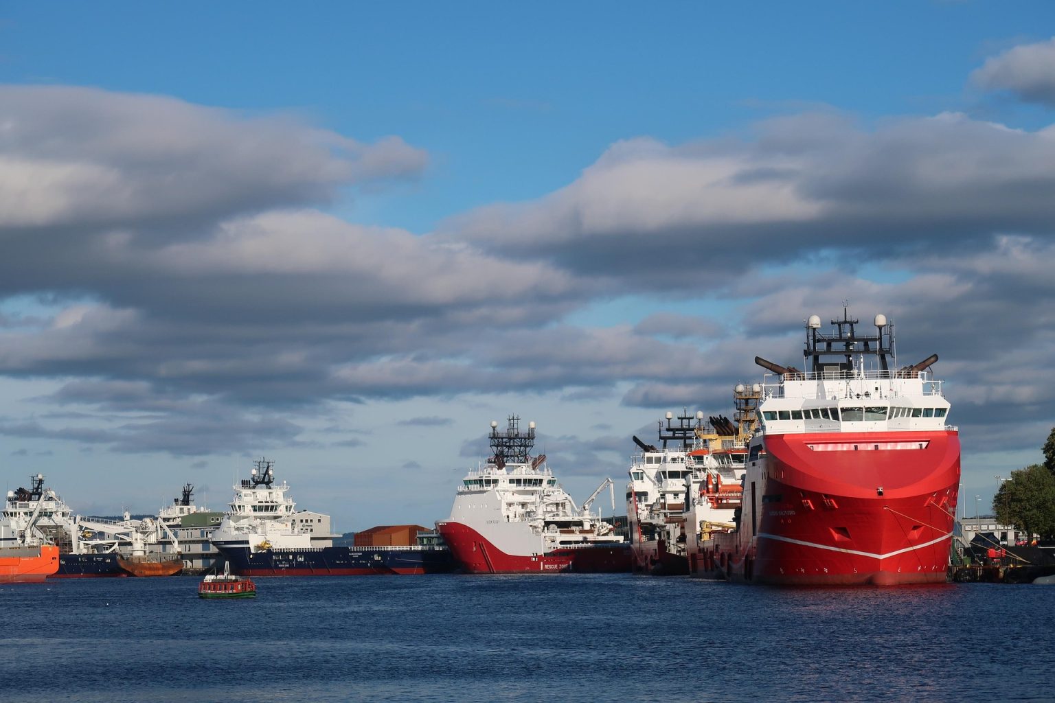 Multiple cargo and supply ships docked at a busy harbor under a partly cloudy sky, showcasing modern maritime shipping operations
