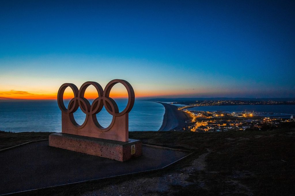 A large stone sculpture of the Olympic rings stands on a cliff overlooking the sea at sunset, with a coastal town illuminated below and a curving shoreline extending into the distance.