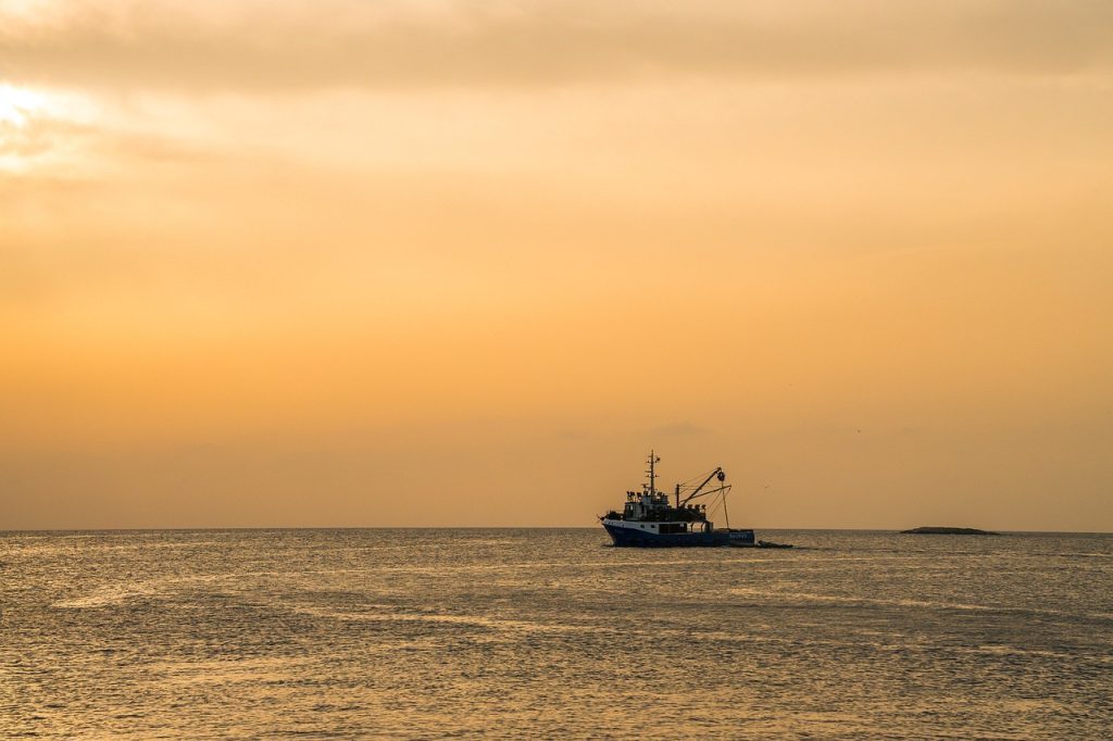 A small fishing boat sails on calm waters under a golden-orange sunset sky, with a distant island barely visible on the horizon.
