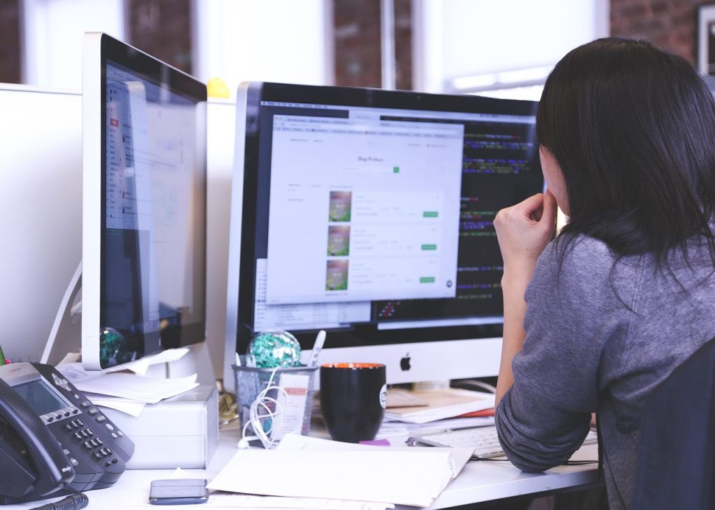 **Alt Text:**
Woman working at a cluttered desk with dual monitors, reviewing code and product listings on a screen in a modern office environment.
