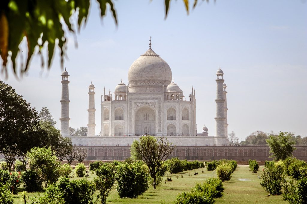 **Alt Text:**
The Taj Mahal, a white marble mausoleum with a large dome and four surrounding minarets, set against a clear blue sky and viewed from a lush green garden in Agra, India.
