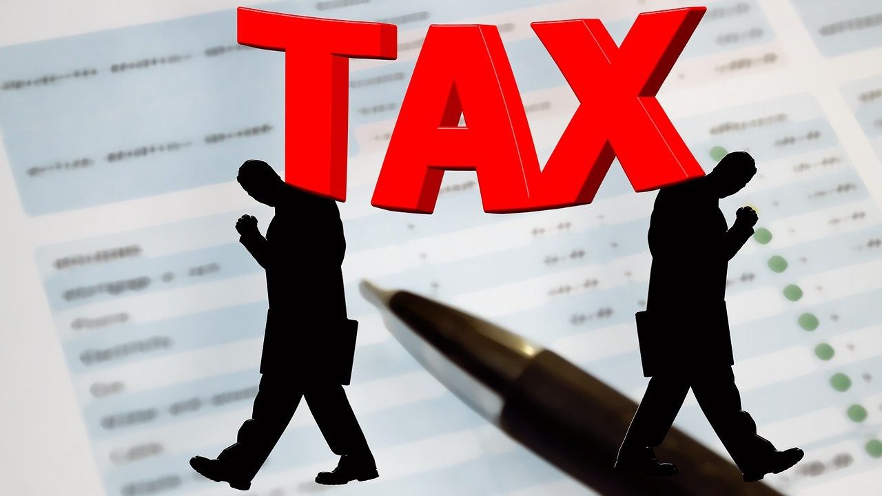 "Two silhouetted figures walking alongside a large pen, with the word 'TAX' prominently displayed in red, set against a blurred background of a budget document."