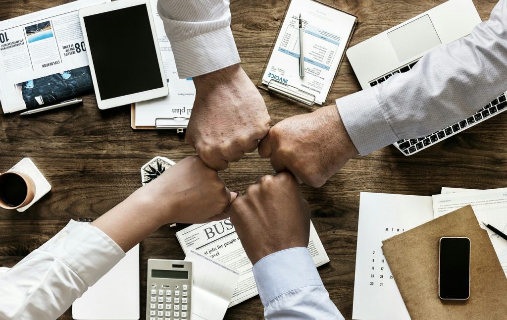Four individuals are seen fist-bumping over a wooden office desk, symbolizing teamwork and collaboration. The desk is cluttered with office supplies including a laptop, a tablet, a calculator, documents labeled “INVOICE” and “BUSINESS,” notebooks, a pen, a smartphone, a coffee cup, and a small potted plant. All individuals are wearing business attire, suggesting a professional setting.