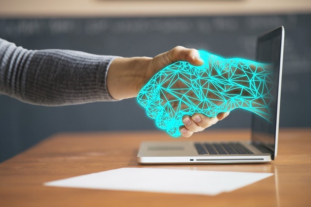 A human hand shaking a glowing digital hand emerging from a laptop screen, symbolizing the connection between technology and humans or digital collaboration.
