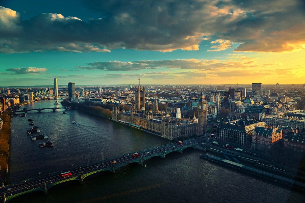 A panoramic sunset view of central London featuring the River Thames, the Palace of Westminster, and the Elizabeth Tower (Big Ben). The Westminster Bridge crosses the river in the foreground, with red double-decker buses visible. The city skyline and dramatic clouds are illuminated by the golden light of the setting sun.