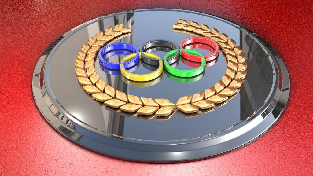 A metallic Olympic emblem featuring the five interlocking colored rings (blue, black, red, yellow, and green) encircled by a golden laurel wreath, set on a reflective silver background with a red textured border.
