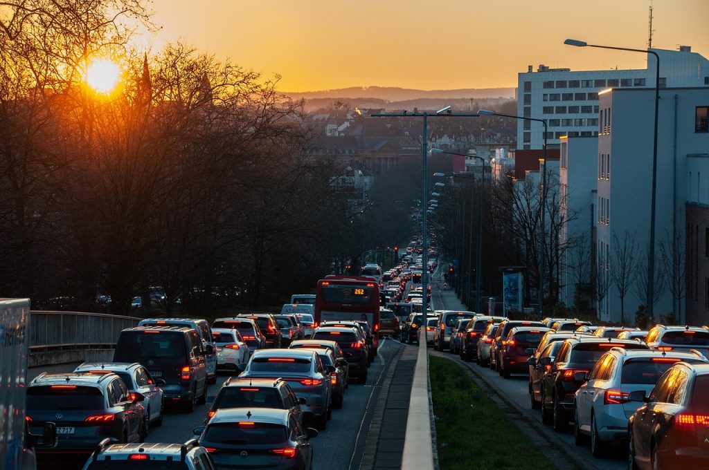 The image shows a traffic jam during sunset in an urban area. Cars are lined up on both sides of a divided road, with a red public bus marked "262" in the center. The sun is setting behind leafless trees, casting a warm glow over the scene. Buildings line the right side of the road, and the horizon shows a hilly silhouette in the background.
