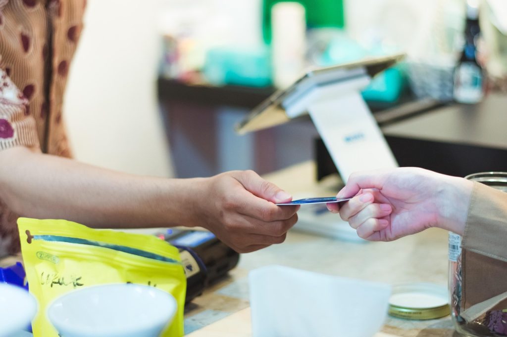 A close-up of a person handing a credit card to a cashier across a store counter with a payment terminal and product packages visible.