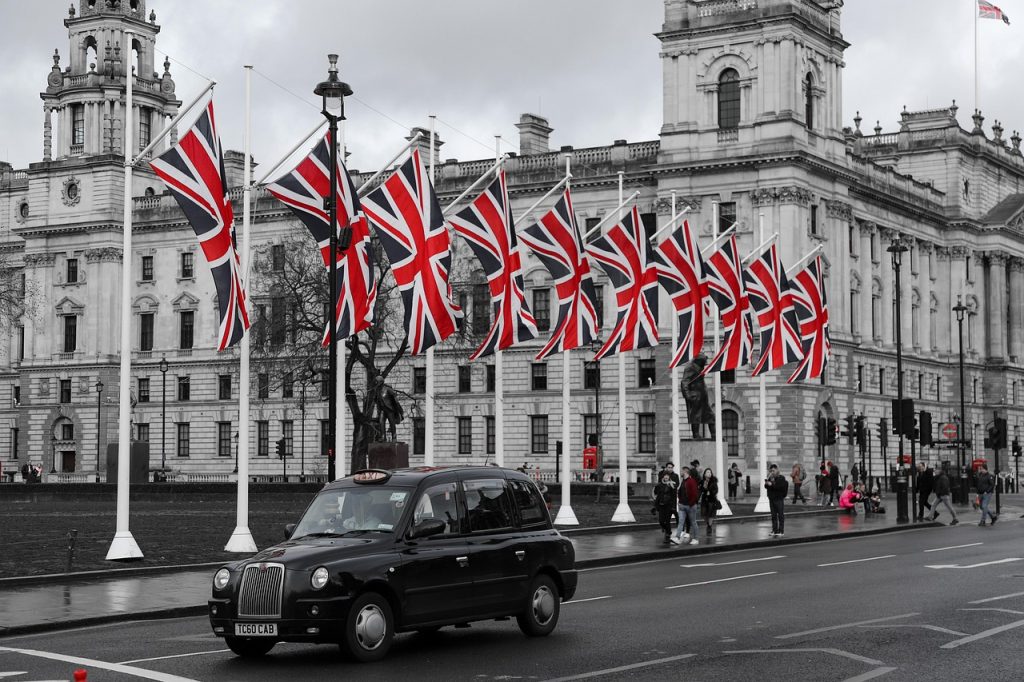 This image appears to be taken in Westminster, London. The scene features numerous Union Jack flags prominently displayed, and a classic black London taxi in the foreground. The architectural style of the building in the background is consistent with the UK government buildings in Whitehall. This combination of elements strongly suggests a national or ceremonial event or display in the area associated with British governance. If you'd like help editing or analyzing something in the image, just let me know!