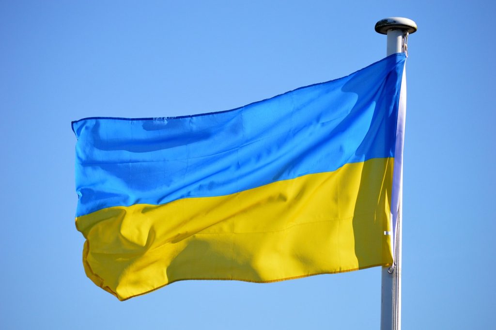 **Alt text:** The national flag of Ukraine with blue and yellow horizontal stripes, flying on a flagpole against a clear blue sky.
