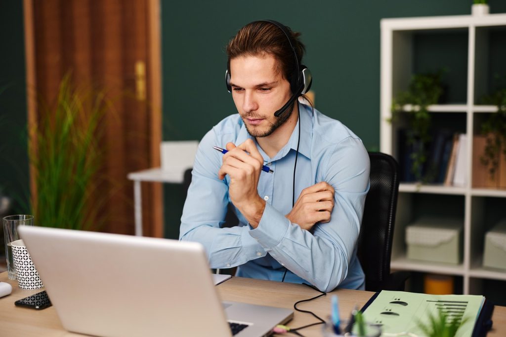 **Alt Text:**
Man in a blue shirt wearing a headset, focused during a video call at a desk with a laptop, documents, and office supplies.
