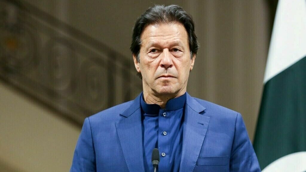 Imran Khan stands in front of a microphone and Pakistan's flag, wearing a serious expression and a blue sherwani, amid the May 9 trial developments.