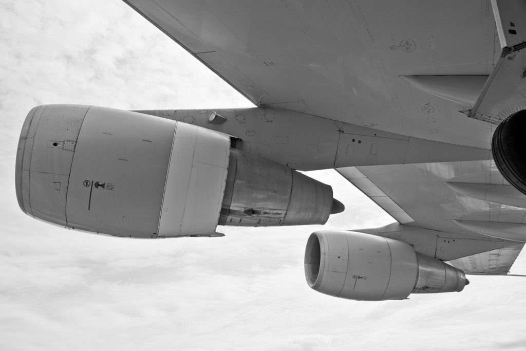 A black and white photograph showing the underside of a large commercial aircraft wing with four jet engines attached. The image is taken from a low angle, capturing the structural details of the engines and wing. The cloudy sky serves as the background.
