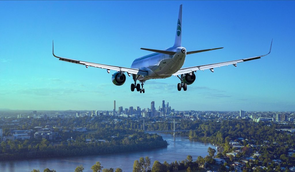A commercial airplane is captured from behind as it descends for landing over a cityscape. The aircraft has its landing gear deployed and is flying over a river with bridges and urban greenery. In the background, a skyline of tall buildings is visible under a clear blue sky.