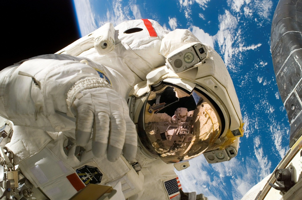 An astronaut in a white space suit is floating in space with Earth visible in the background. The astronaut’s reflective visor shows a mirrored image of another astronaut and part of the spacecraft. Various equipment and patches are visible on the suit, and the blue and white planet below adds dramatic contrast to the scene.